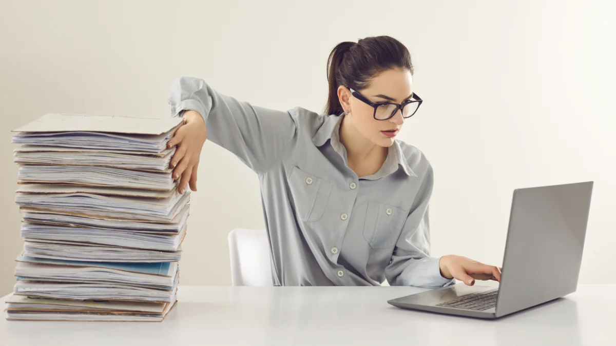 Bookkeeper working through a tall stack of paperwork at a laptop